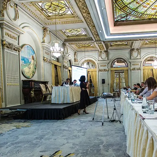 Lectoră prezentând în picioare, lângă scenă, sub plafonul ornamentat al sălii Le Diplomate