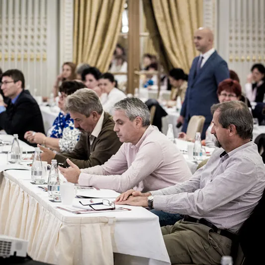 Participanți atenți la mese, cu mape și pixuri, urmărind prezentarea