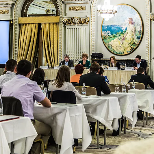 Prezidiul conferinței la masa din fața sălii, sub tabloul rotund, cu participanții văzuți din spate